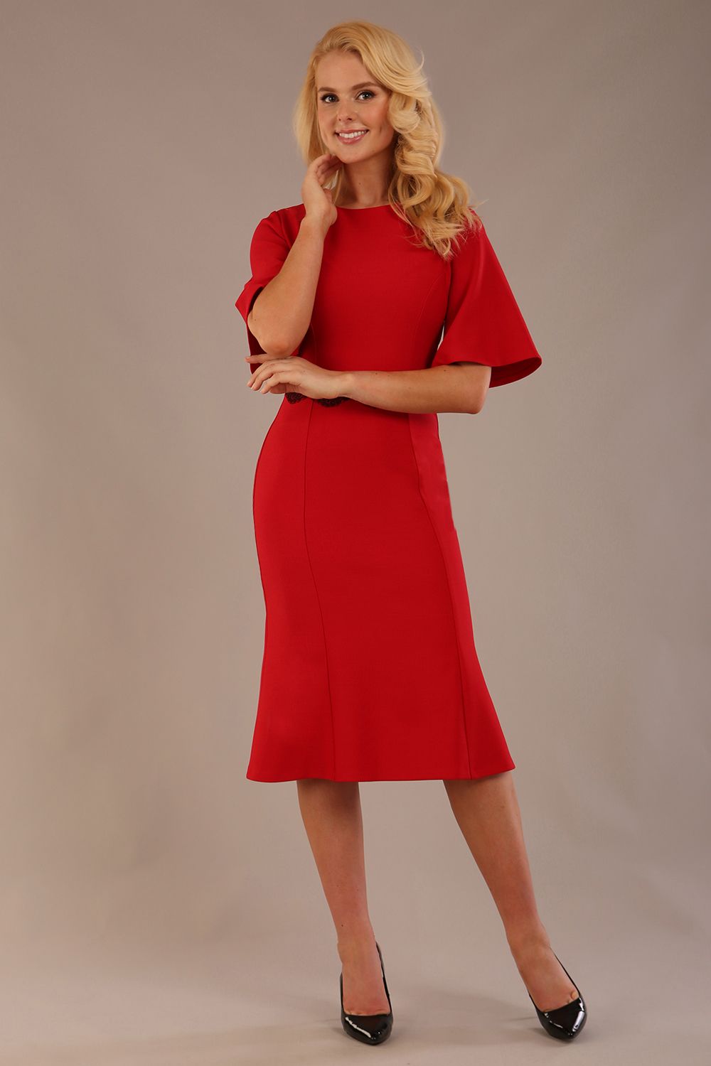 Model wearing the Lovell dress in salsa red, showcasing the fluted sleeves and black lace waistband against a beige background