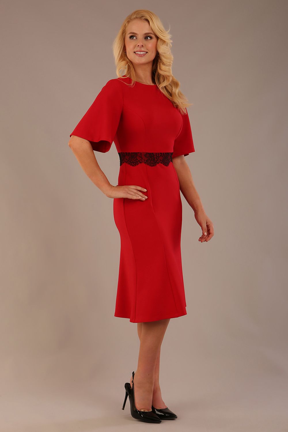 Model wearing the Lovell dress in salsa red, showcasing the fluted sleeves and black lace waistband against a beige background