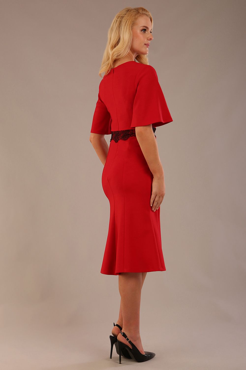 Model wearing the Lovell dress in salsa red, showcasing the fluted sleeves and black lace waistband against a beige background back view