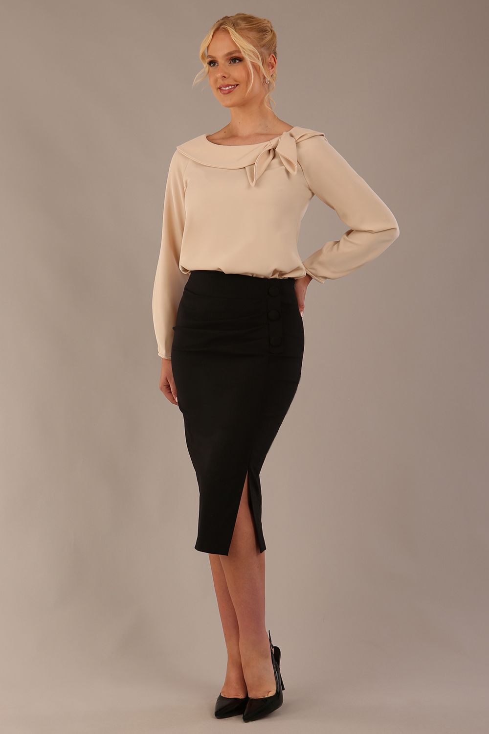 model wearing a Cressida long sleeve top in almond cream, with a boat neckline and asymmetric bow, styled with a black skirt front view