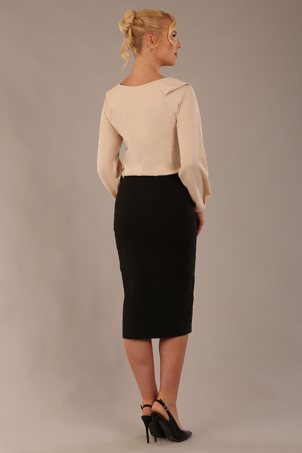 model wearing a Cressida long sleeve top in almond cream, with a boat neckline and asymmetric bow, styled with a black skirt back view