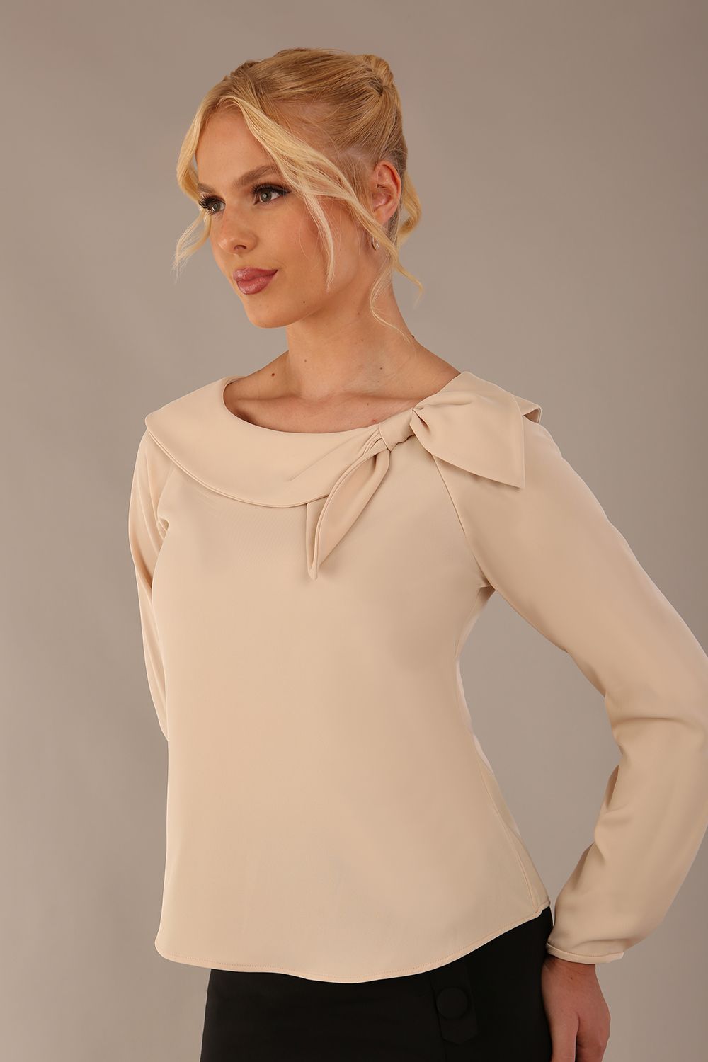 model wearing a Cressida long sleeve top in almond cream, with a boat neckline and asymmetric bow, styled with a black skirt close up view