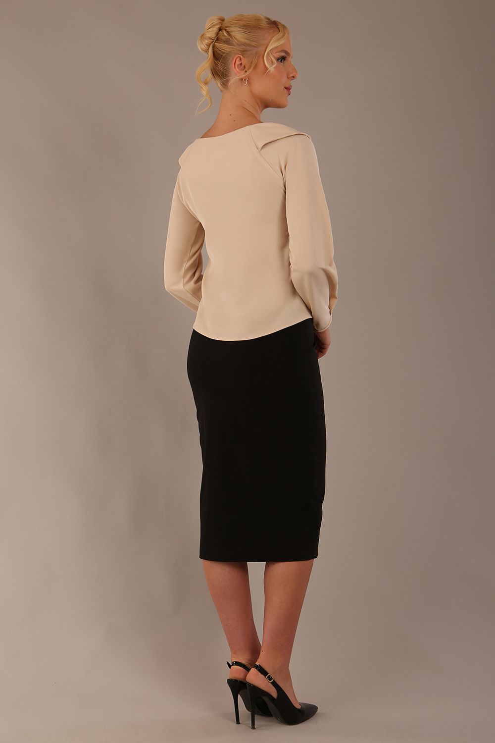 model wearing a Cressida long sleeve top in almond cream, with a boat neckline and asymmetric bow, styled with a black skirt back view