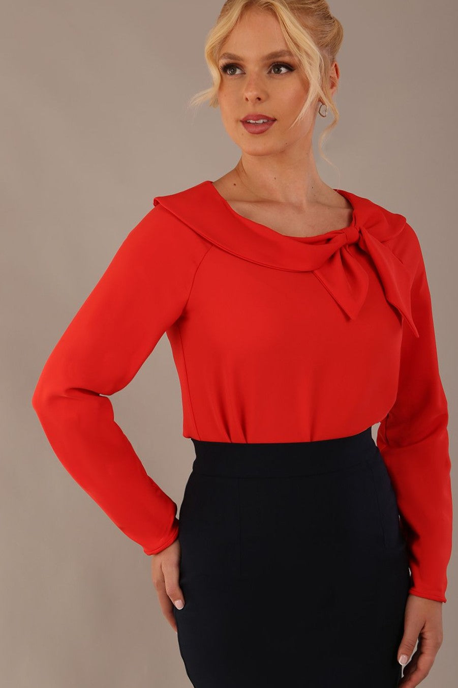 model wearing a Cressida long sleeve top in poppy red, with a boat neckline and asymmetric bow, styled with a pencil skirt close up