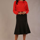 Brunette model wearing a Cressida long sleeve top in poppy red, with a boat neckline and asymmetric bow, styled with a black fishtail skirt.