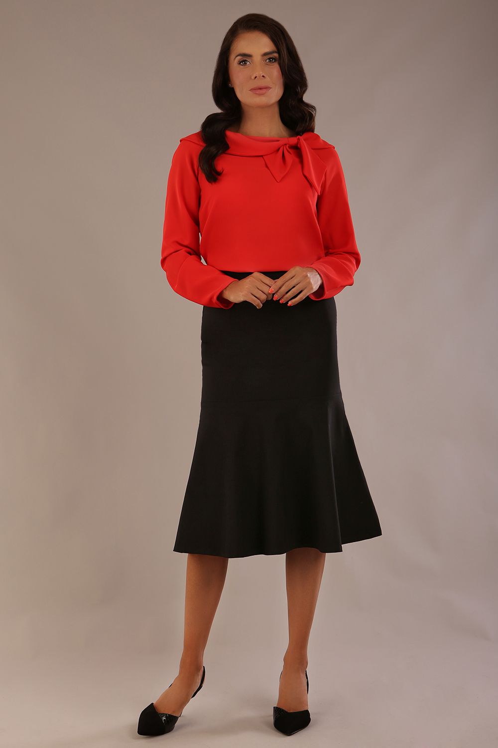 Brunette model wearing a Cressida long sleeve top in poppy red, with a boat neckline and asymmetric bow, styled with a black fishtail skirt.