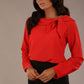 Brunette model wearing a Cressida long sleeve top in poppy red, with a boat neckline and asymmetric bow, styled with a black fishtail skirt.