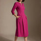 Woman wearing a diva catwalk Amanda Pleated a-line dress in a magenta pink colour with pockets against a beige background (Magenta)