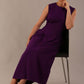 Model wearing the Cleo pleated midaxi dress in purple with hands in the discreet side pockets sitting on a chair against a plain background