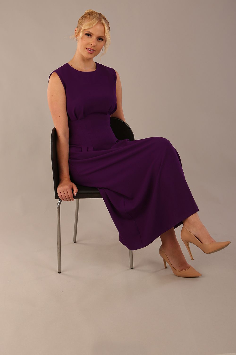 Model wearing the Cleo pleated midaxi dress in purple with hands in the discreet side pockets sitting on a chair against a plain background