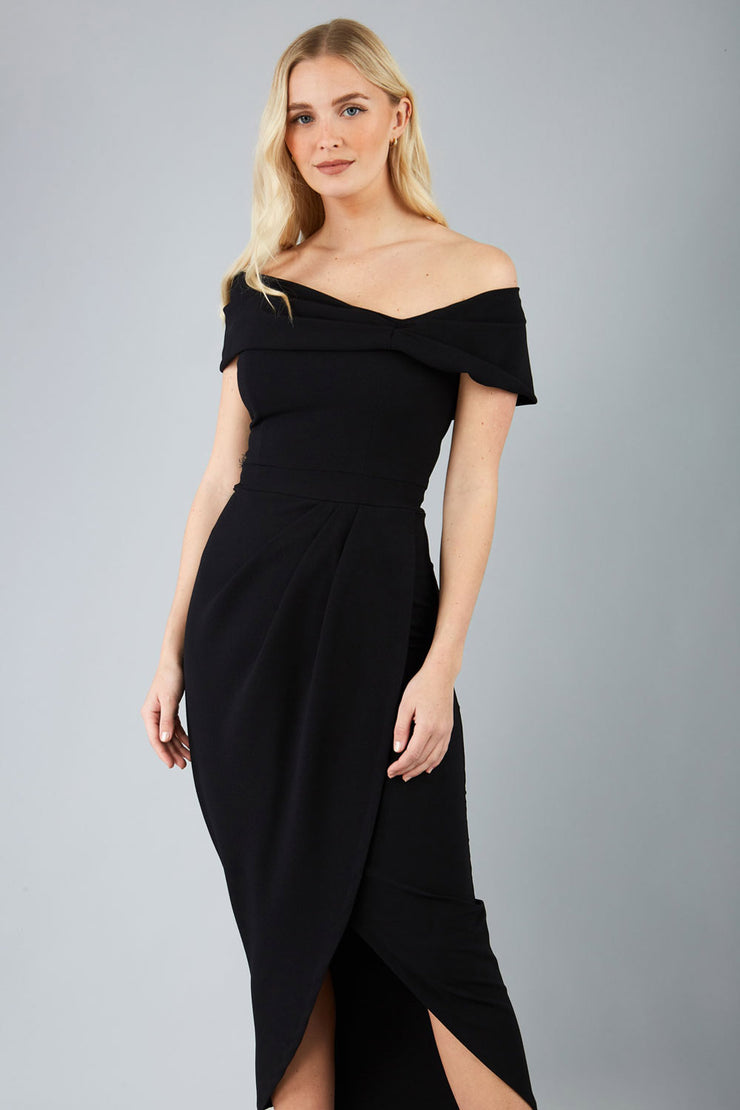 odel wearing diva catwalk vegas calf length black midaxi dress with wide bardot neckline and open shoulders with a large opening at the front of the skirt with pleating coming down long skirt front