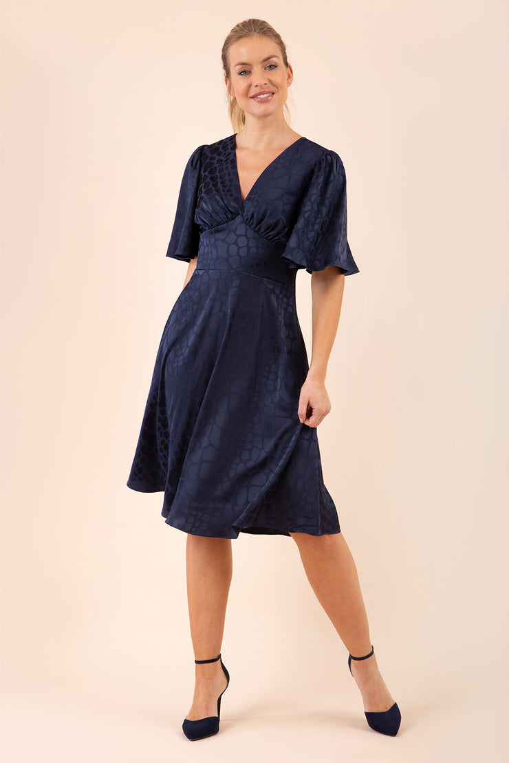 brunette model wearing diva catwalk cambrian satin swing dress with bell short sleeve wide asymmetric waistband and pleating under bust and low v-neckline in navy front