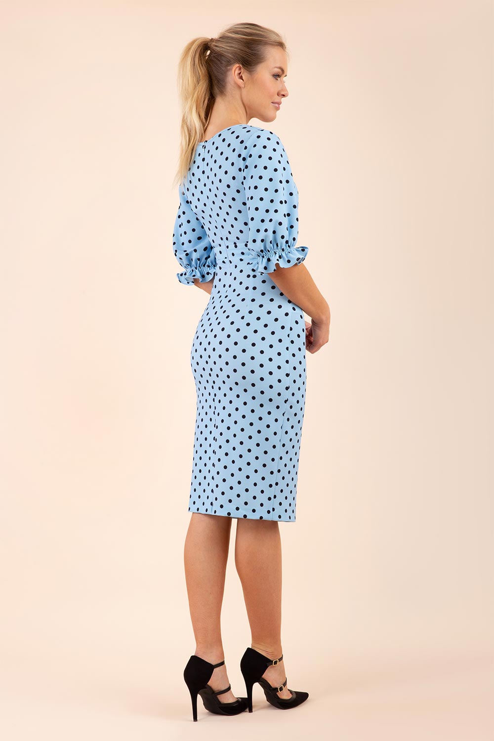 Blonde model wearing Diva Catwalk Palacio Pencil dress v neckline and pleating across the tummy with puffed short sleeves in blue polka dot back