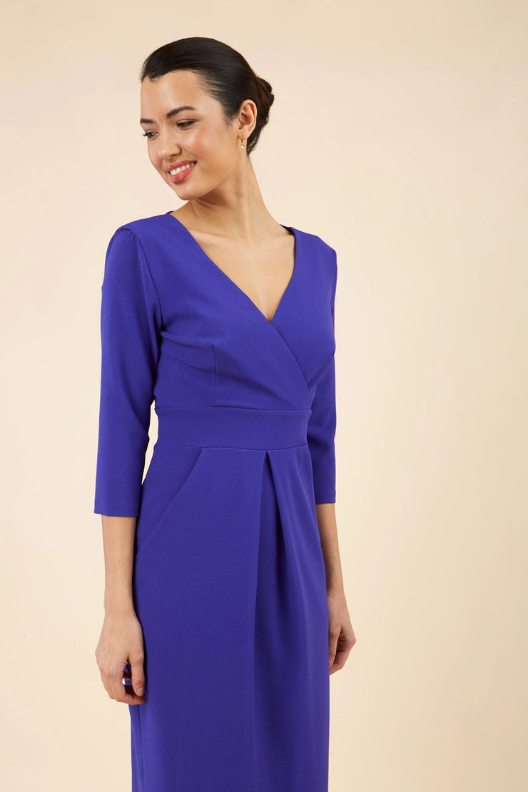 brunette model wearing diva catwalk wimpole a-line flattering dress with lowered v-neck and three quarter sleeve and cinching waistband and widened skirt with pleating in spectrum indigo front