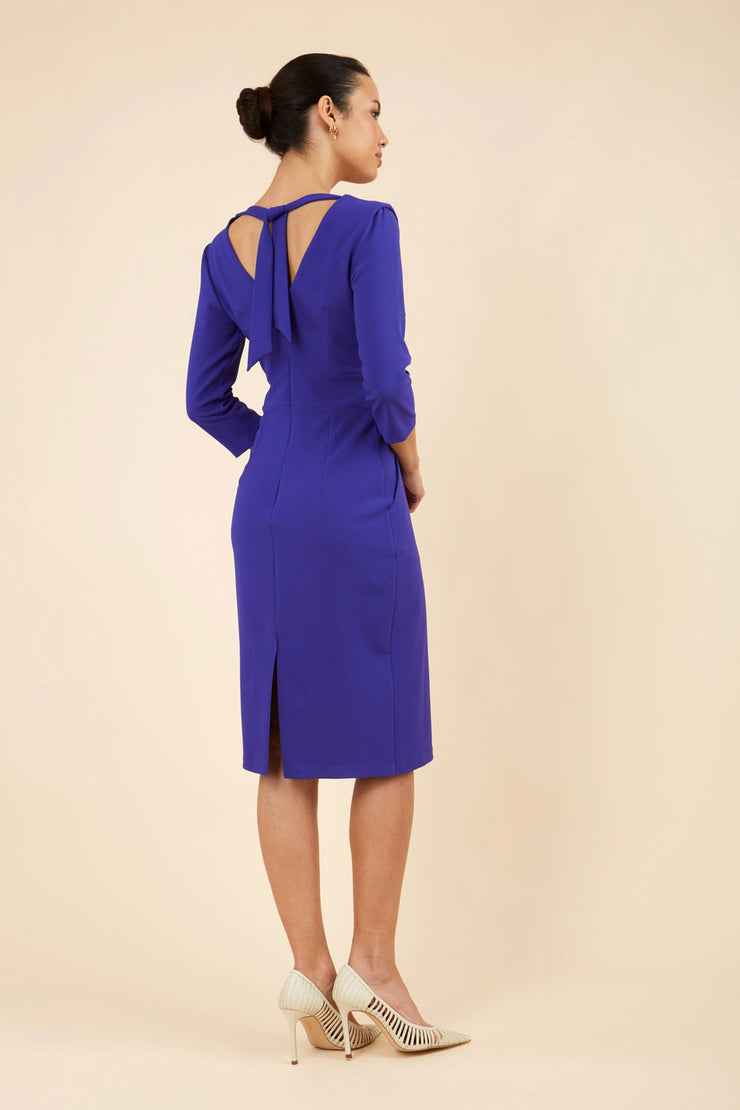 brunette model wearing diva catwalk wimpole a-line flattering dress with lowered v-neck at the back and tie detail and three quarter sleeve and and widened skirt with vent  in spectrum indigo back