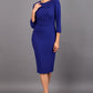 brunette model wearing diva catwalk elodie pencil fitted dress with rounded neckline and button detail and three quarter sleeve with pleating across the body from side triangle detail in oxford blue front
