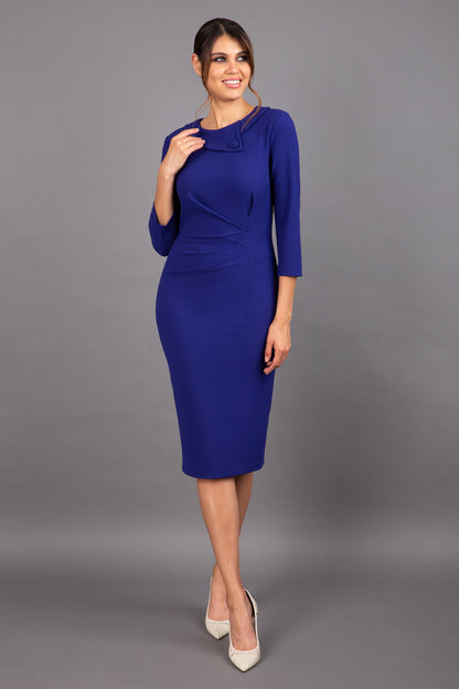 brunette model wearing diva catwalk elodie pencil fitted dress with rounded neckline and button detail and three quarter sleeve with pleating across the body from side triangle detail in oxford blue front
