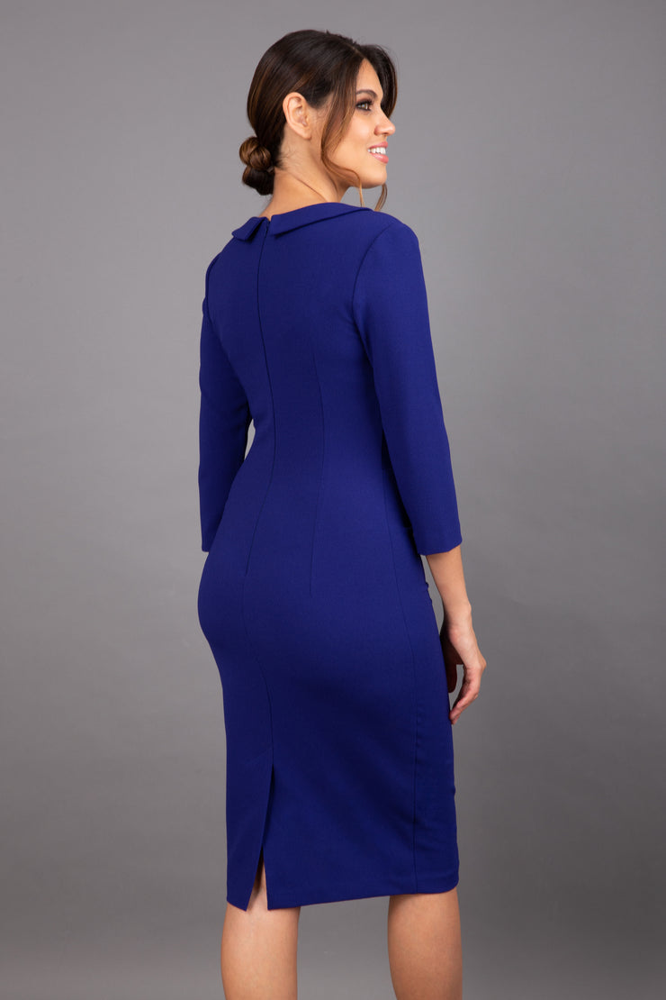 brunette model wearing diva catwalk elodie pencil fitted dress with rounded neckline and button detail and three quarter sleeve with pleating across the body from side triangle detail in oxford blue back