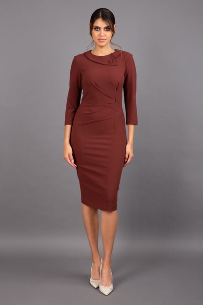 brunette model wearing diva catwalk elodie pencil fitted dress with rounded neckline and button detail and three quarter sleeve with pleating across the body from side triangle detail in mahogany front