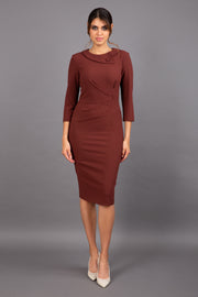 brunette model wearing diva catwalk elodie pencil fitted dress with rounded neckline and button detail and three quarter sleeve with pleating across the body from side triangle detail in mahogany front