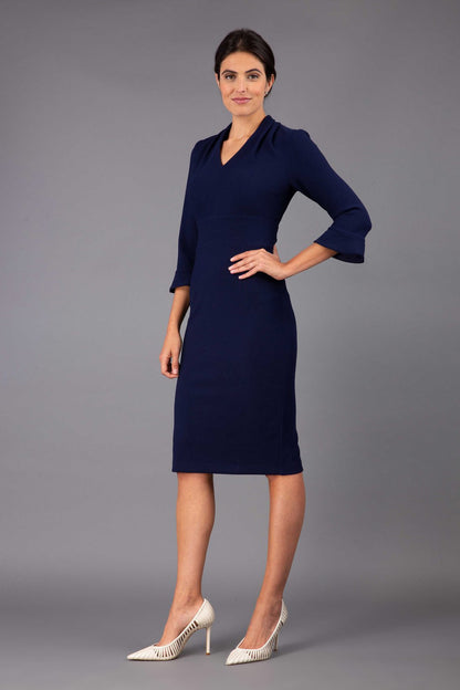 brunette model wearing diva catwalk adriana pencil dress with low v-neck and pleating around shoulders with three quarter sleeve detailed on end and wide waistband in dark navy front