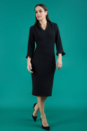 brunette model wearing diva catwalk adriana pencil dress with low v-neck and pleating around shoulders with three quarter sleeve detailed on end and wide waistband in black front