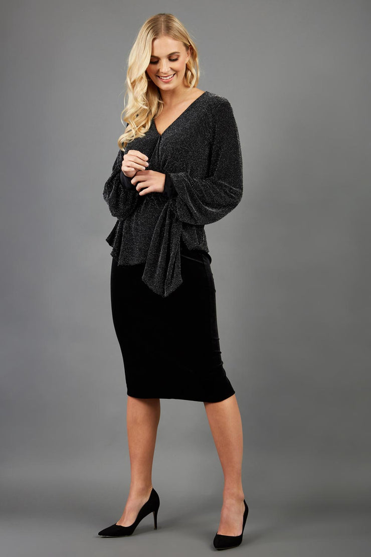 Blonde hair model is wearing a jersey sparkle v neck wrap blouse with bishop sleeve detail and bow on a side front image