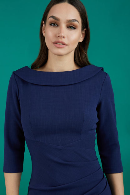 A brunette model is wearing a round neckline pencil dress with pleating on the tummy area in navy front image