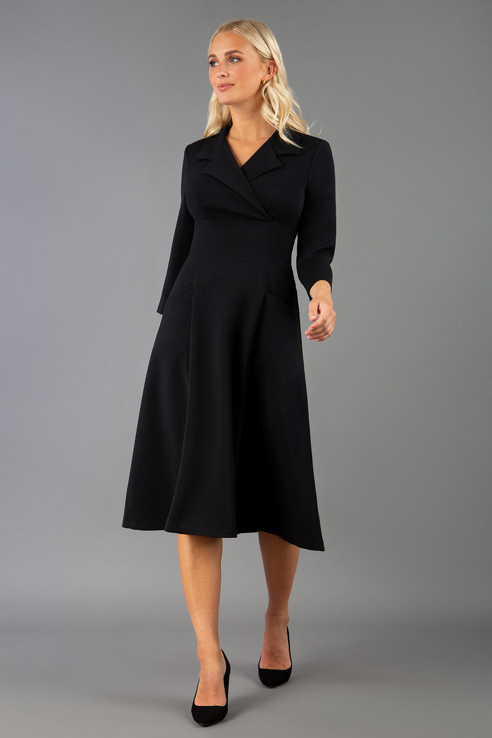 A blonde model is wearing a swing black three quarter sleeve dress with oversized collar and pockets in the skirt  